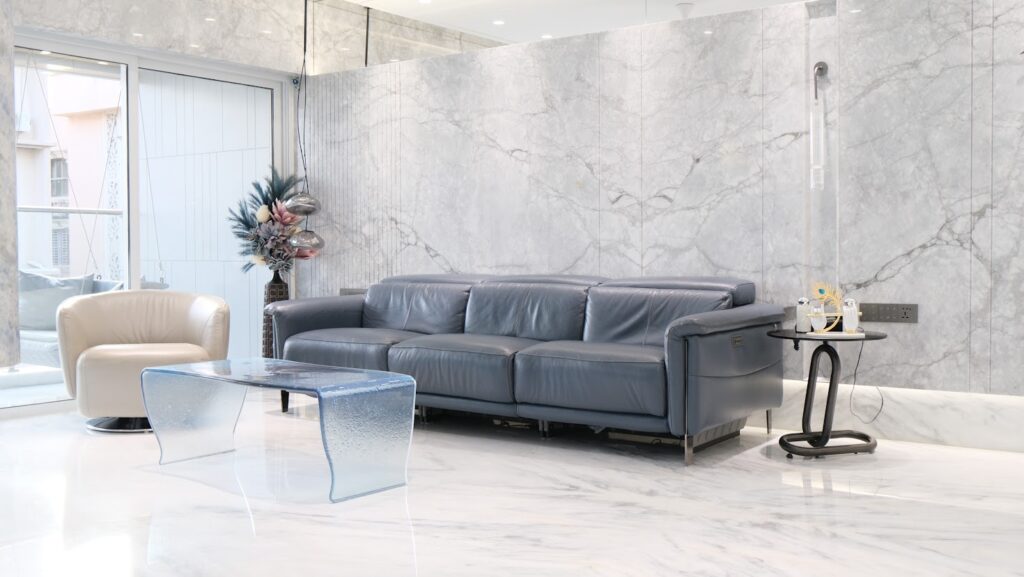 Modern living room featuring a blue leather sofa, white armchair, glass coffee table, and marble walls.